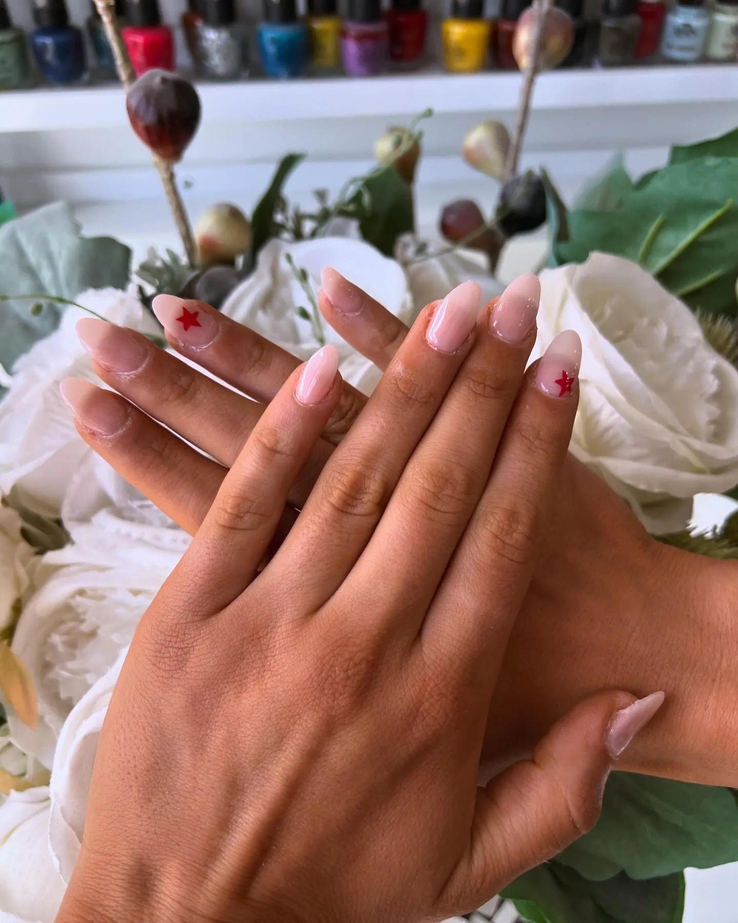 Short almond nails in milky pink with a single hand-painted red star on two accent nails, staged on white roses below a polish shelf