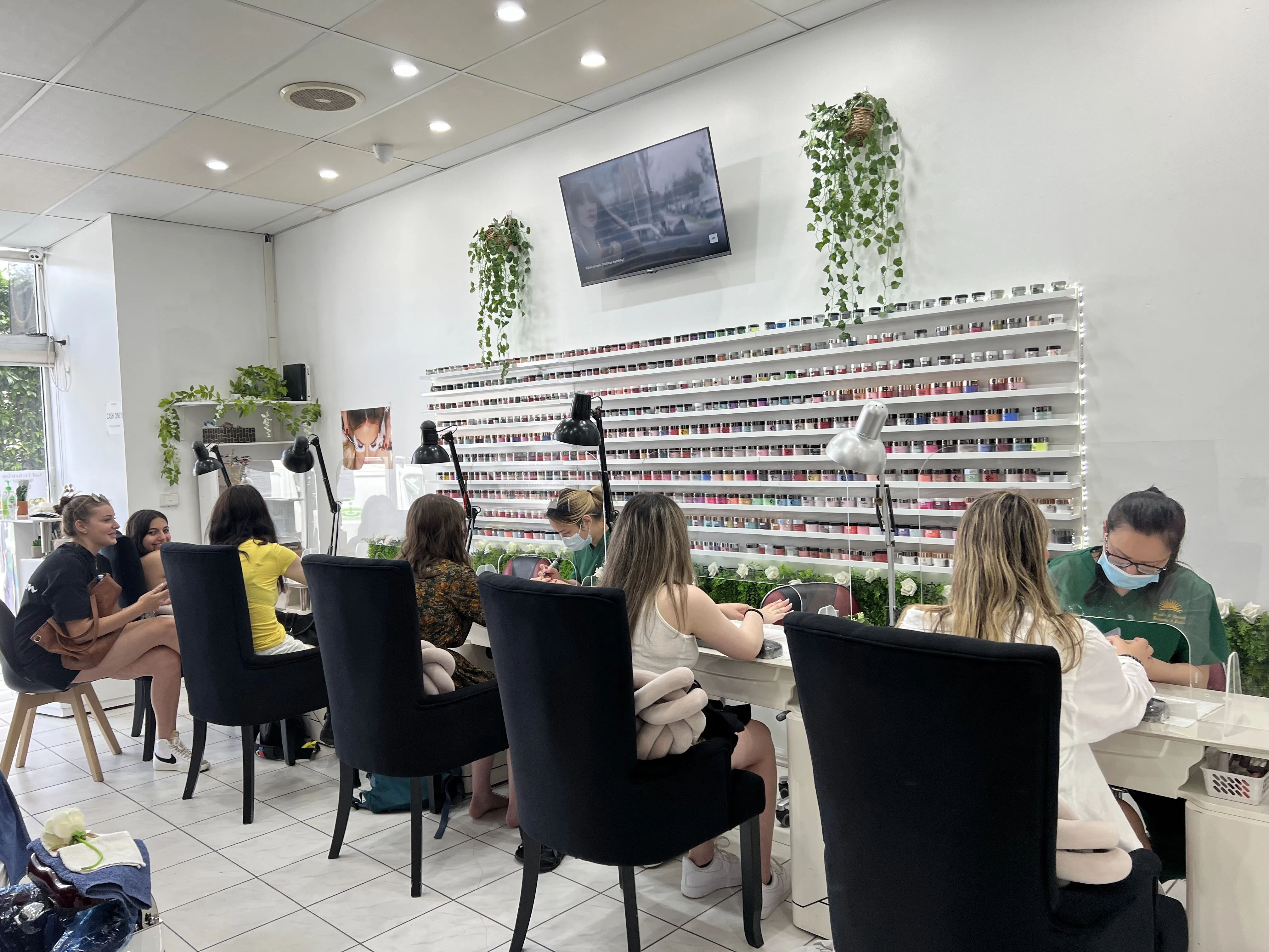 Sun Nails & Beauty at work — eight clients being served, two technicians in forest-green sunburst-logo polos, the SNS colour wall behind them