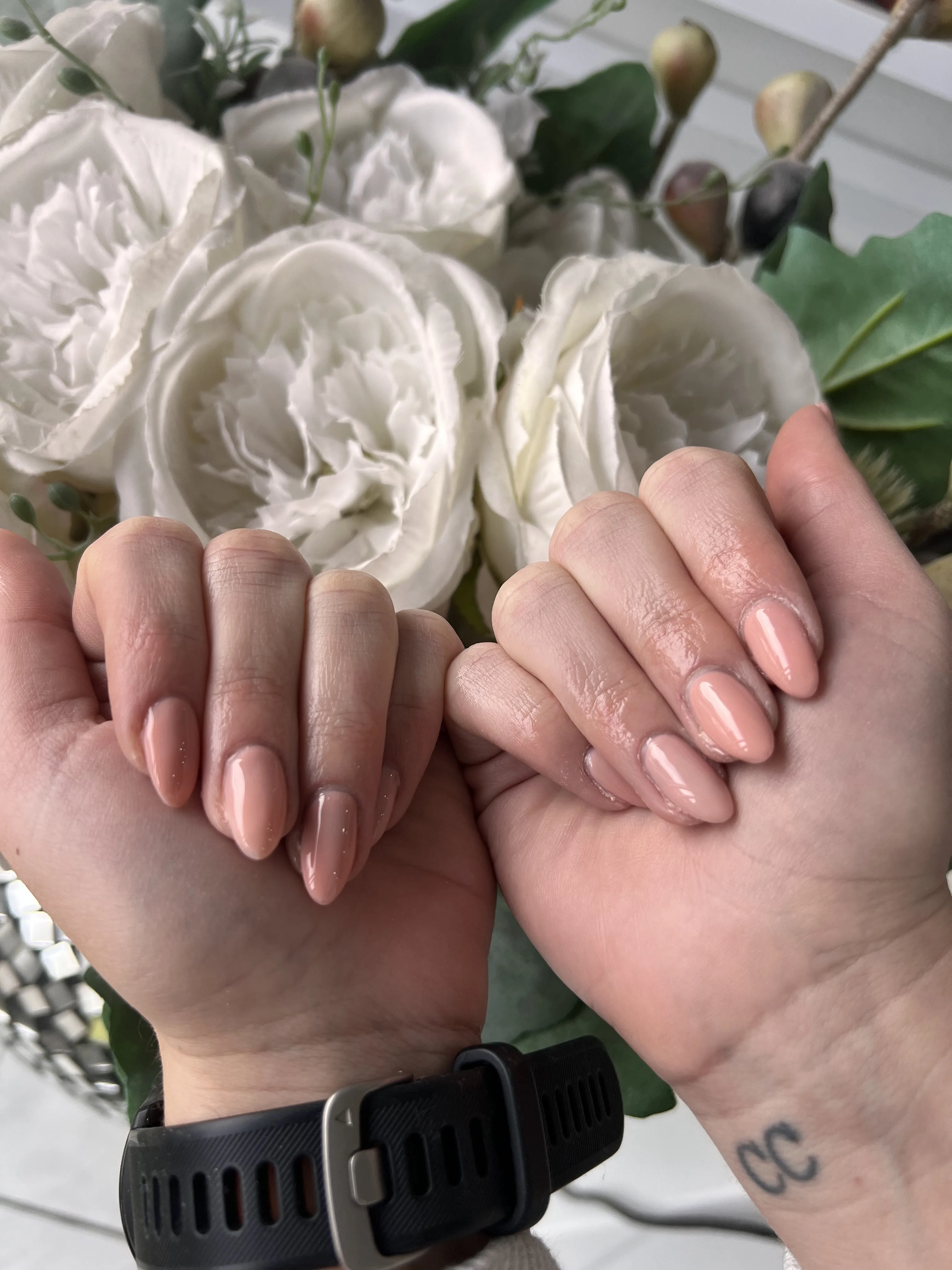 Medium almond nails in a warm nude-peach gloss, two hands clasped, white roses behind