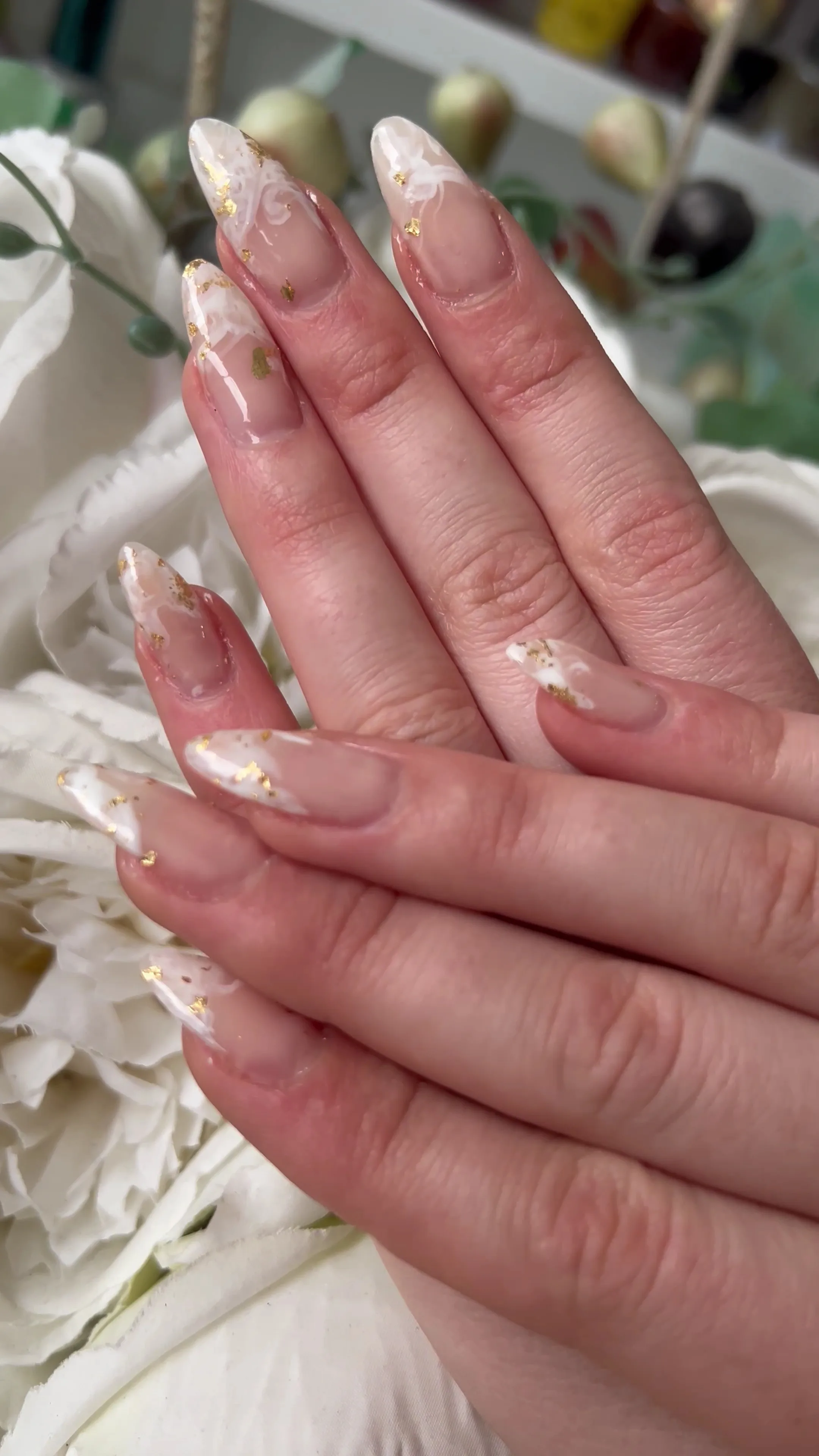 Long almond nails — sheer milky base with white marble swirls and scattered gold leaf, staged on white roses