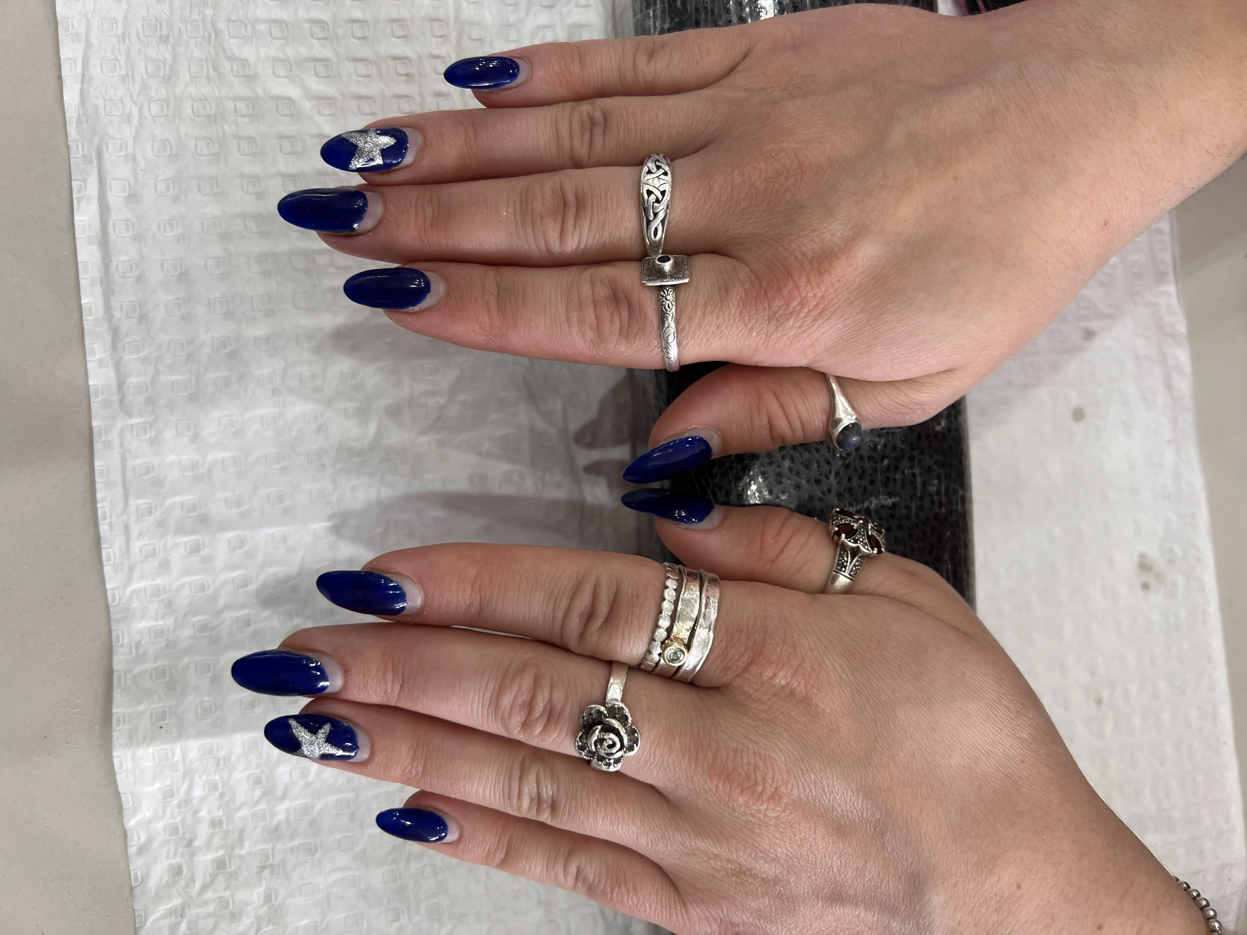 Medium almond nails in deep gloss navy with a hand-painted silver star on accent nails, hands stacked with ornate silver rings